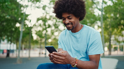 Fototapeta na wymiar Closeup portrait of young African American man in light blue t-shirt sitting on city park bench and using his smartphone. Man looks at photos, videos in his mobile phone. lifestyle concept.