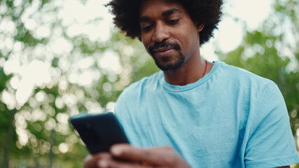 Fototapeta premium Closeup portrait of young African American man in light blue t-shirt sitting on city park bench and using his smartphone. Man looks at photos, videos in his mobile phone. lifestyle concept.