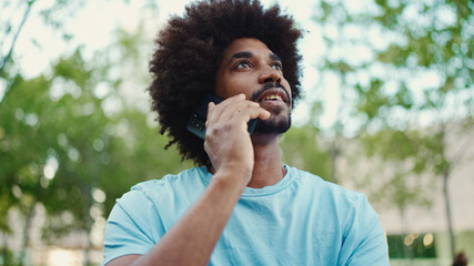 Closeup young African American man in light blue t-shirt sitting on city park bench and talking on...
