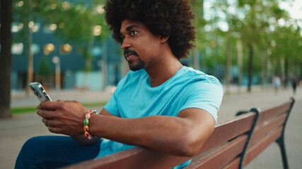 Close-up of young African American man in blue t-shirt sitting on park bench and talking on...