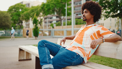 Close-up of young African American man wearing shirt sitting relaxed on park bench looking away....