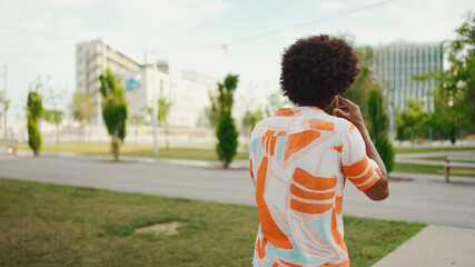 Close-up of young African American man wearing shirt walking and talking on smartphone on path in the park. Man emotionally talking on mobile phone. Lifestyle concept.