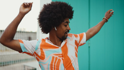 Close-up of cheerful young African American man wearing shirt listening to music in wireless headphones and dancing on light blue wall background. Lifestyle concept.