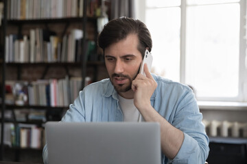 Serious millennial businessman sit at workplace looks at laptop while talks on smartphone, speak to client, provide assistance remotely by phonecall. Modern tech, business, workflow, multitask concept