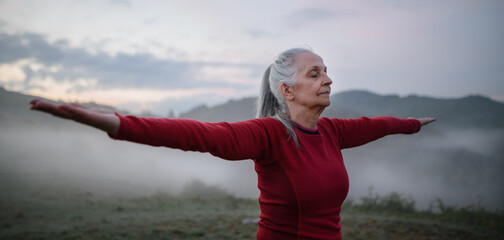 Fototapeta na wymiar Senior woman doing breathing exercise in nature on early morning with fog and mountains in background.