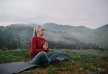 Naklejka na ściany i meble Senior woman doing breathing exercise in nature on early morning with fog and mountains in background.