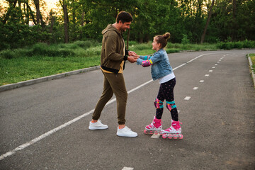A father teaching his daughter roller skating in a park on summer day.