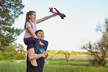 Fototapeta premium Cute girl riding on father's shoulder playing with toy airplane