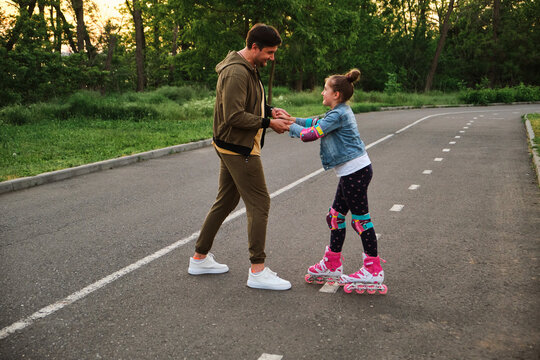 A Father Teaching His Daughter Roller Skating In A Park On Summer Day.