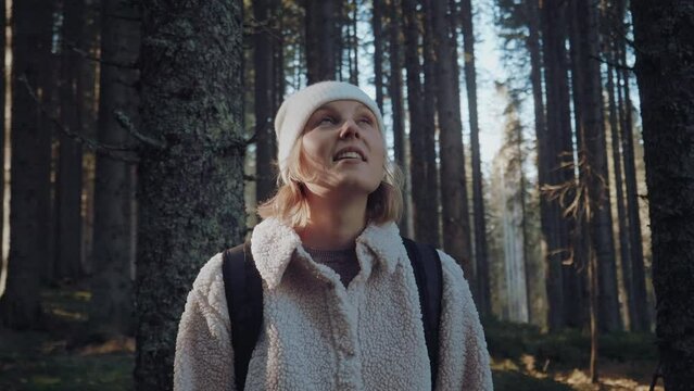 Close up shot of a young woman with blonde hair and backpack hiking in untouched green forest in autumn. The morning sun is making flares. Active healthy female is smiling.
