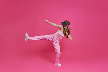 Playful young woman in virtual reality headset imitating fight while standing against pink background