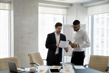 Two multi ethnic businessmen speaking in office, consider documents, share information, planning...