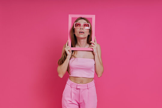 Curious young woman looking through a picture frame while standing against pink background