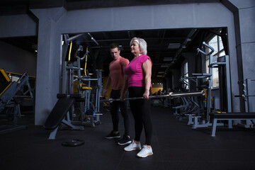 Fototapeta na wymiar Full length shot of an elderly woman lifting barbell, working out with personal trainer at the gym