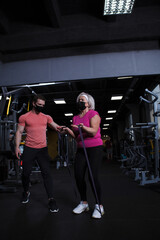 Fototapeta na wymiar Vertical full length shot of a senior woman and her fintess coach wearing medical face masks at the gym
