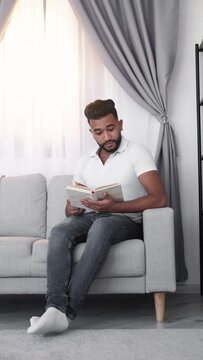 Home relax. Casual man. Book lover. Arabian guy sitting comfortable sofa and reading in light room interior vertical.