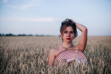 A young, slender, in black panties in a field of wheat covers her chest with a straw hat.