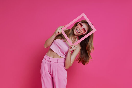 Happy young woman looking through a picture frame while standing against pink background