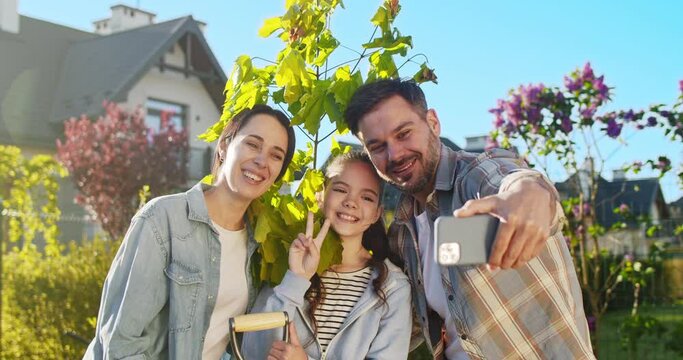 Happy Caucasian family in garden taking selfie photo with smartphone on sunny summer day. Handsome father making picture with wife and teen daughter in orchard with mobile phone camera.