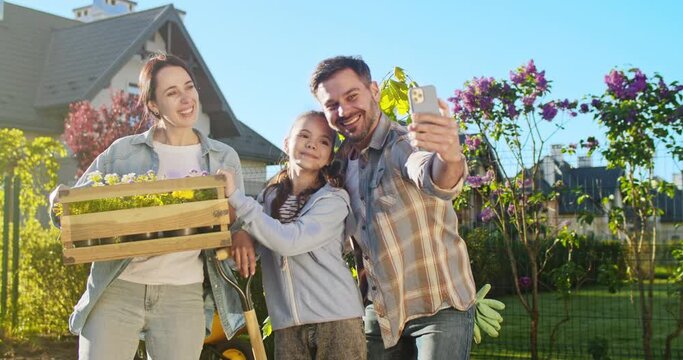 Happy family in garden taking selfie photo on smartphone on sunny summer day. Holding box with flowers seedlings. Handsome father making picture with wife and teen daughter in orchard on mobile phone.
