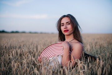 A young, slender, in black panties in a field of wheat covers her chest with a straw hat.