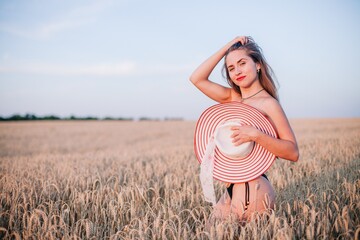 A young, slender, in black panties in a field of wheat covers her chest with a straw hat.