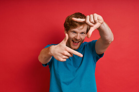 Playful redhead man gesturing picture frame and smiling while standing against red background