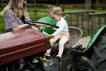 baby girl is sitting on retro tractor, toddler changes gears, the mother in summer dress watches over her daughter while she is playing - obrazy, fototapety, plakaty