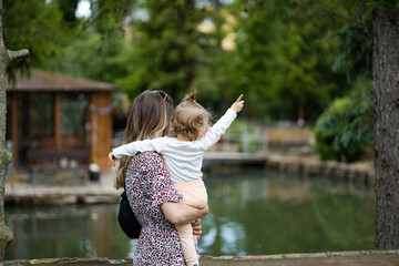 mother in a summer dress holding her daughter in hands, toddler is pointing upwards, country water in the background - obrazy, fototapety, plakaty