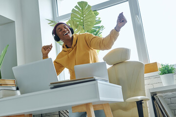 Happy mixed race man in headphones dancing while standing near working place