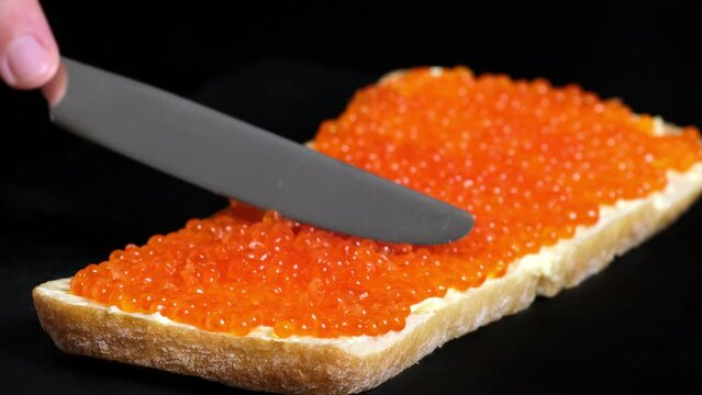 Spreads red caviar on bread with knife. Chef smears salmon caviar on toast. Close-up of knife is spreading caviar on bread. Tasting salmon caviar salted roe, black background slow motion