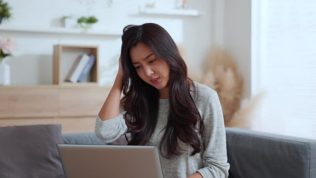 Asian businesswoman works at computer from home Young woman entrepreneur upset stressed typing at the laptop reading bad news on social media. Irritability and numbness scratching the head.