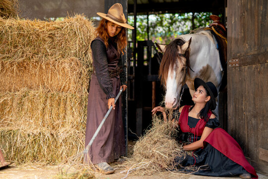 Two Asian woman with cowgirl costume enjoy to feed dry grasses to horse near stable in their farm.