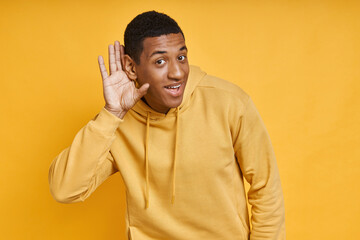 Curious mixed race man holding hand near ear while standing against yellow background