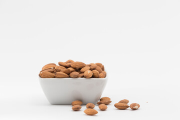view of a square bowl containing a bunch of peeled almonds, placed on a white background, one almond has fallen out of the bowl