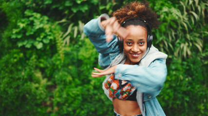 Closeup, smiling African girl wearing denim jacket, in crop top with national pattern walks in city park listening to music on headphones and dancing on green trees background.