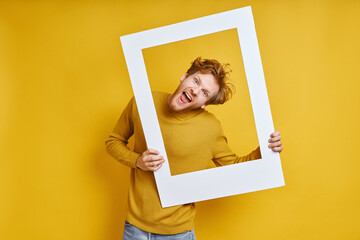 Playful redhead man looking through a picture frame while standing against yellow background