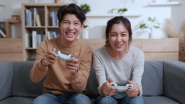 Happy millennia couple asian sitting on the couch playing video games, using joystick controllers. Woman and man in love have fun playing in video games console at home together.