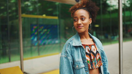 Cute African girl with ponytail, wearing denim jacket, in crop top with national pattern, standing at the bus stop and smiling - obrazy, fototapety, plakaty