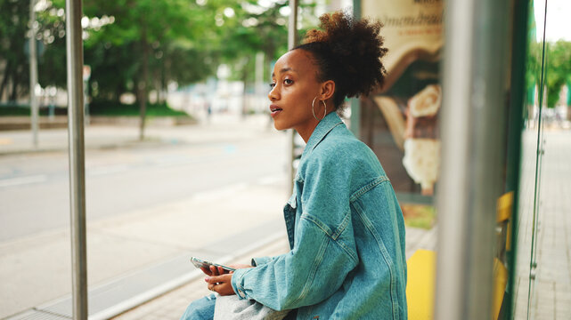 Cute African girl with ponytail, wearing denim jacket, in crop top with national pattern, sitting at the bus stop and using her mobile phone.