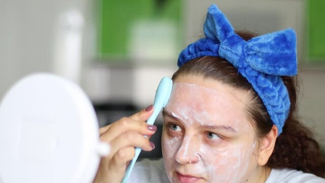 Woman in hairband using brush to apply moisture anti-acne and pimple clay mask, moisturizer cream, looking in mirror. Face skincare, professional product cosmetics. Hygiene and dermatology, massage