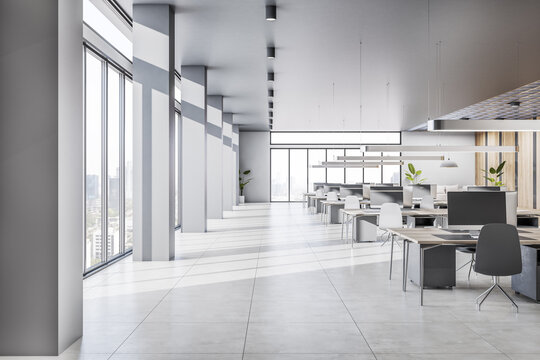 Spacious sunlit coworking office with wooden work tables, concrete floor, grey stylish lamps on light ceiling and city view from panoramic windows. 3D rendering