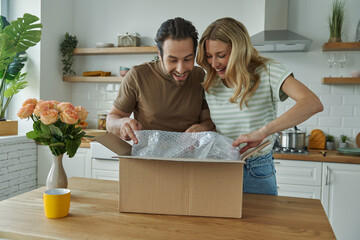 Curious young couple unpacking box while standing at the domestic kitchen - obrazy, fototapety, plakaty