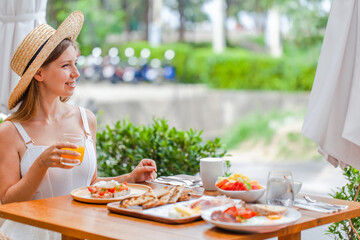 Young smiling woman on breakfast in outdoor restaurant in hotel. Female traveler in straw hat drinking orange juice in modern cafe. Summer vacation