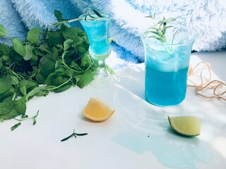 Blue cold drink in a glass with ice cubes on white desk with blue background. Also used mint leaves and rosemary, rope, spoon  with natual sunlight from the open window