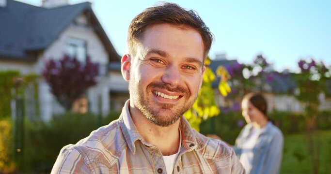 Portrait shot of handsome Caucasian young man standing in garden, looking straight and smiling to camera. Woman and daughter working in orchard on background. Outdoor. Close up.