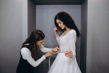 dressmaker helping the bride to put her wedding dress on in clothes shop.