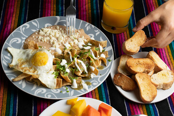 traditional mexican food called chilaquiles in green sauce, with beans, slices of bolillo, tropical fruit of the region: mango, papaya and orange juice, on a typical mexican cloth tablecloth.