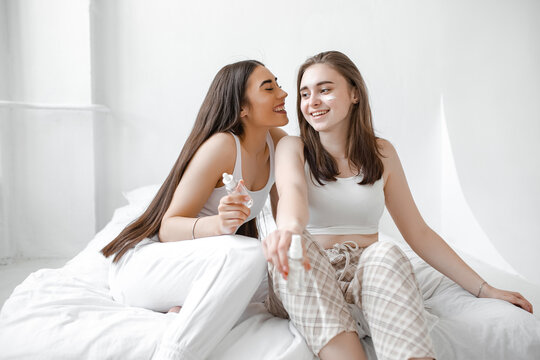 Two young beautiful girls with cosmetics sit on a white bed. Women use serum, cream and facial lotion to take care of their skin. Girlfriends in home spa rest and relax