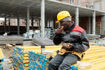 Hungry mature black man in hardhat and workwear eating sandwich and drinking hot tea while sitting...